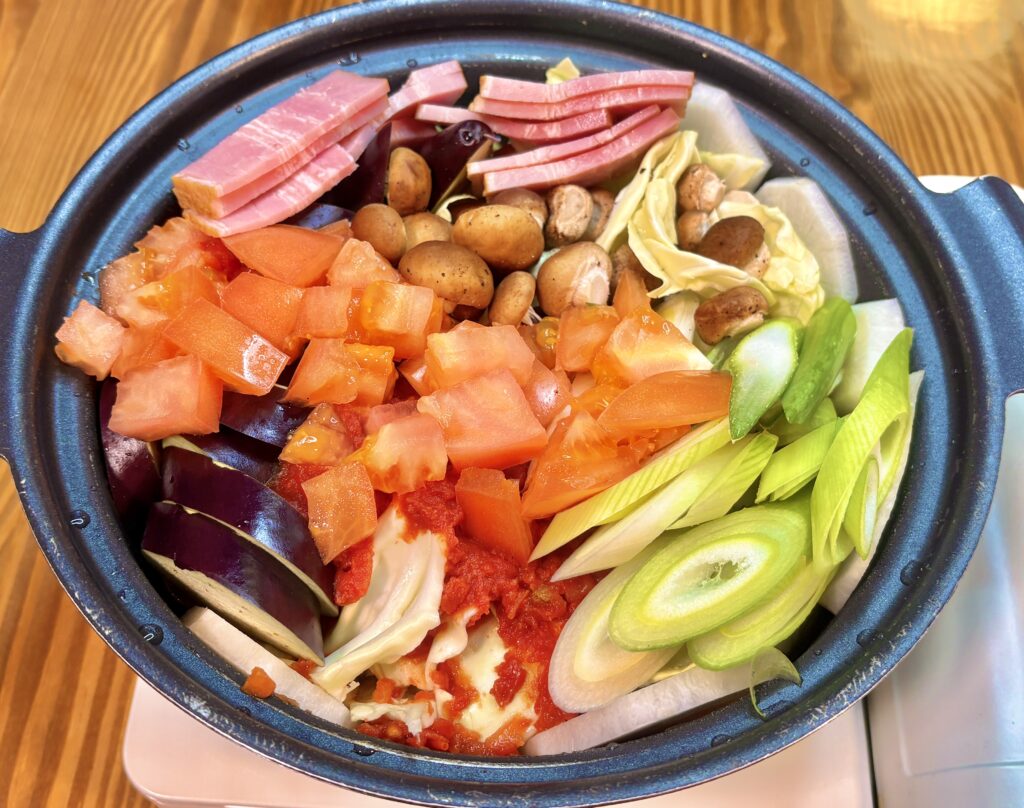 和風トマト鍋の具材を鍋に並べた様子。角切りトマト、なす、キャベツ、長ねぎ、しめじ、ベーコン、などが彩りよく入っている。
