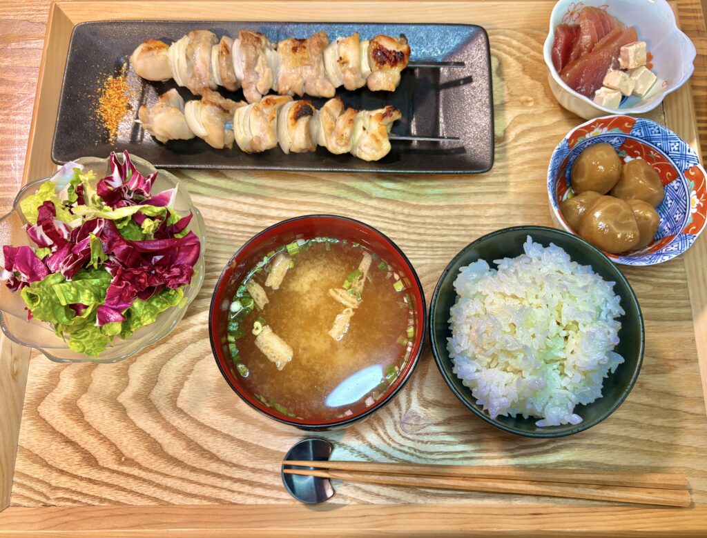 木製トレーの上に、串打ちした焼き鳥2本を黒い長皿に盛り付け、ご飯、油揚げ入りの味噌汁、葉物野菜のサラダ、たまこんにゃく、クリームチーズとキハダマグロの出汁醤油漬けを並べた、家庭的な和定食の全体写真。
