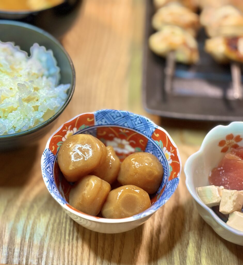色付きの和柄小鉢に盛られた、照りのあるたまこんにゃくのクローズアップ写真。背景にご飯茶碗と焼き鳥がぼんやり写り、温かい食卓の雰囲気が伝わる。
