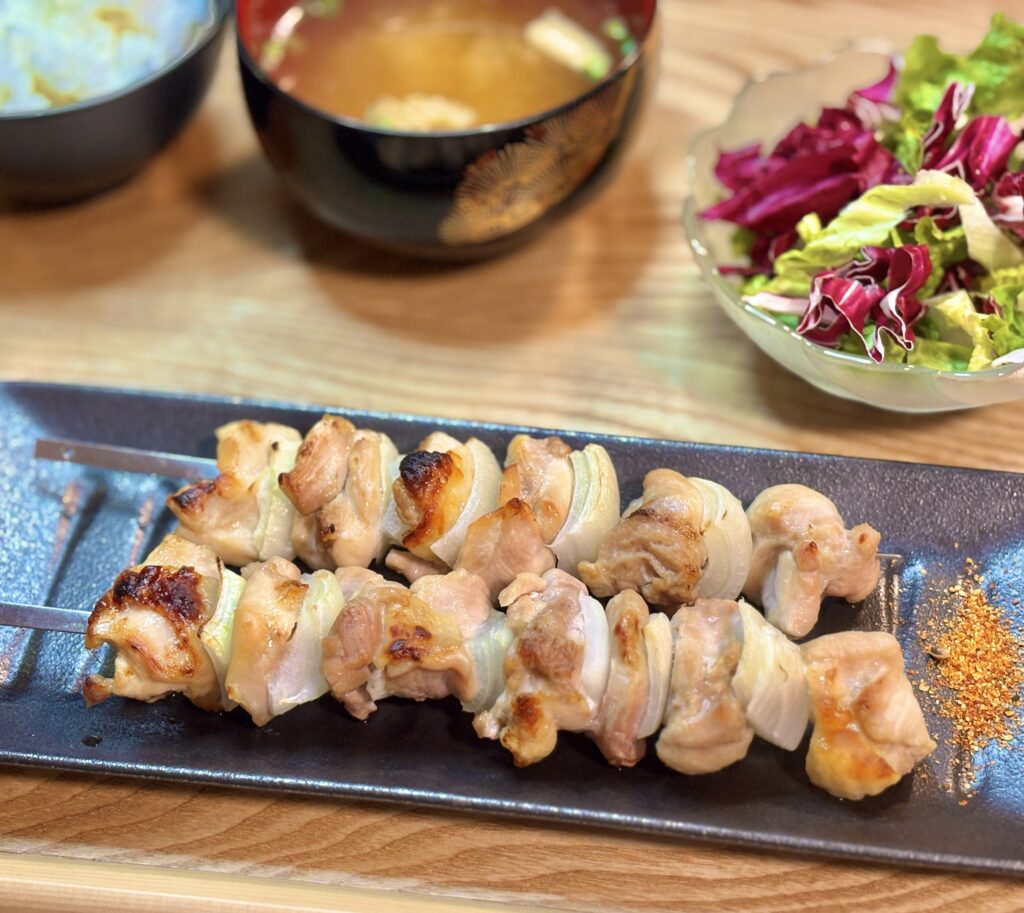 玉ねぎと鶏肉を交互に串打ちした焼き鳥を、黒い長皿にのせたクローズアップ写真。焼き目が香ばしく、横に七味唐辛子が添えられている。
