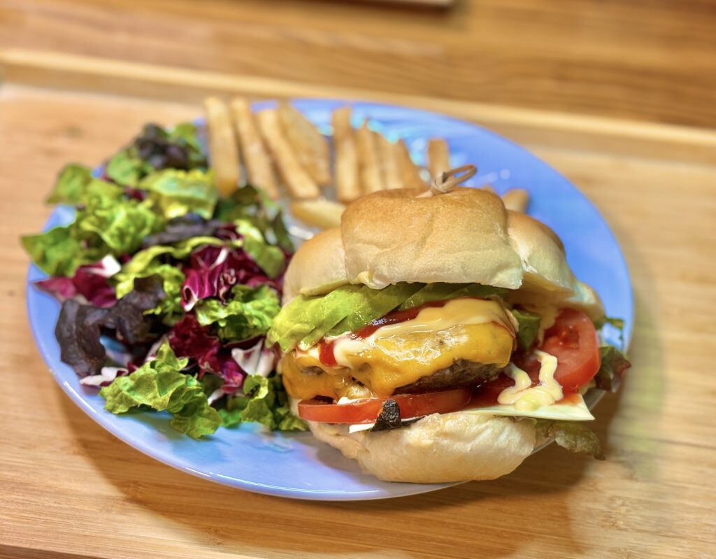 アボカドとチェダーチーズを重ねた手作りハンバーガーが水色の皿に盛られている。
横にはミックスリーフのサラダと細切りフライドポテトが添えられ、木製トレーの上に置かれている。