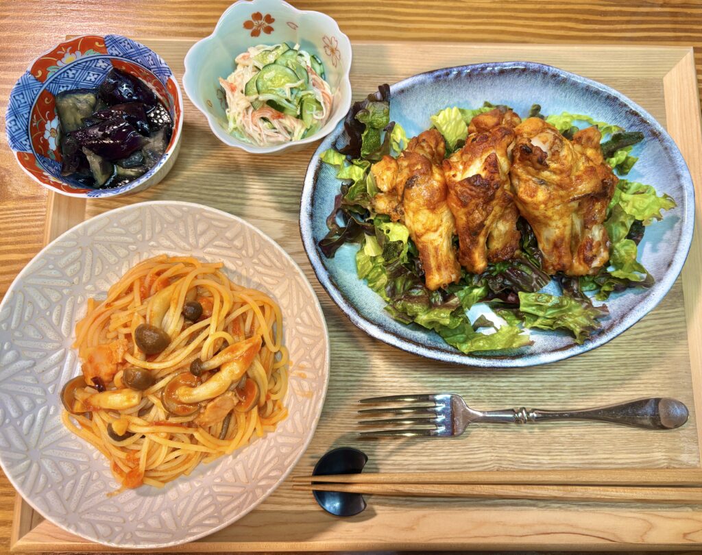 木製トレーの上に並んだ夕食プレート。レタスを敷いたタンドリーチキン、しめじとベーコンのガーリックトマトパスタ、カニカマときゅうりの和え物、茄子の煮浸しが彩りよく盛り付けられている。