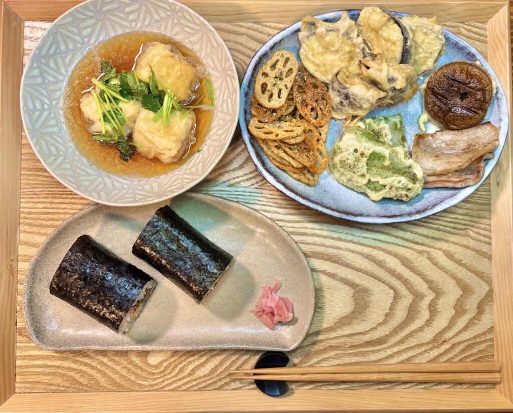 木製のお盆の上に節分の食卓が並んでいる。買ってきた太巻き寿司が半分に切られて器に盛られ、横に紅しょうがが添えられている。別の器にはレンコンチップス、野菜の天ぷら、しいたけの素揚げ、なすやピーマンの揚げ物が彩りよく並び、奥には三つ葉をのせた揚げ出し豆腐が盛られている。
