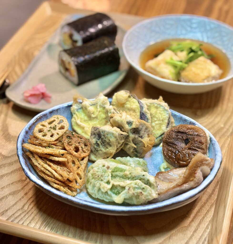 器いっぱいに盛られた野菜の揚げ物。薄切りレンコンのチップス、なすやピーマンの天ぷら、しいたけの素揚げなどが並び、衣の色合いや野菜の形がはっきり分かる。奥には太巻き寿司と揚げ出し豆腐が見える。
