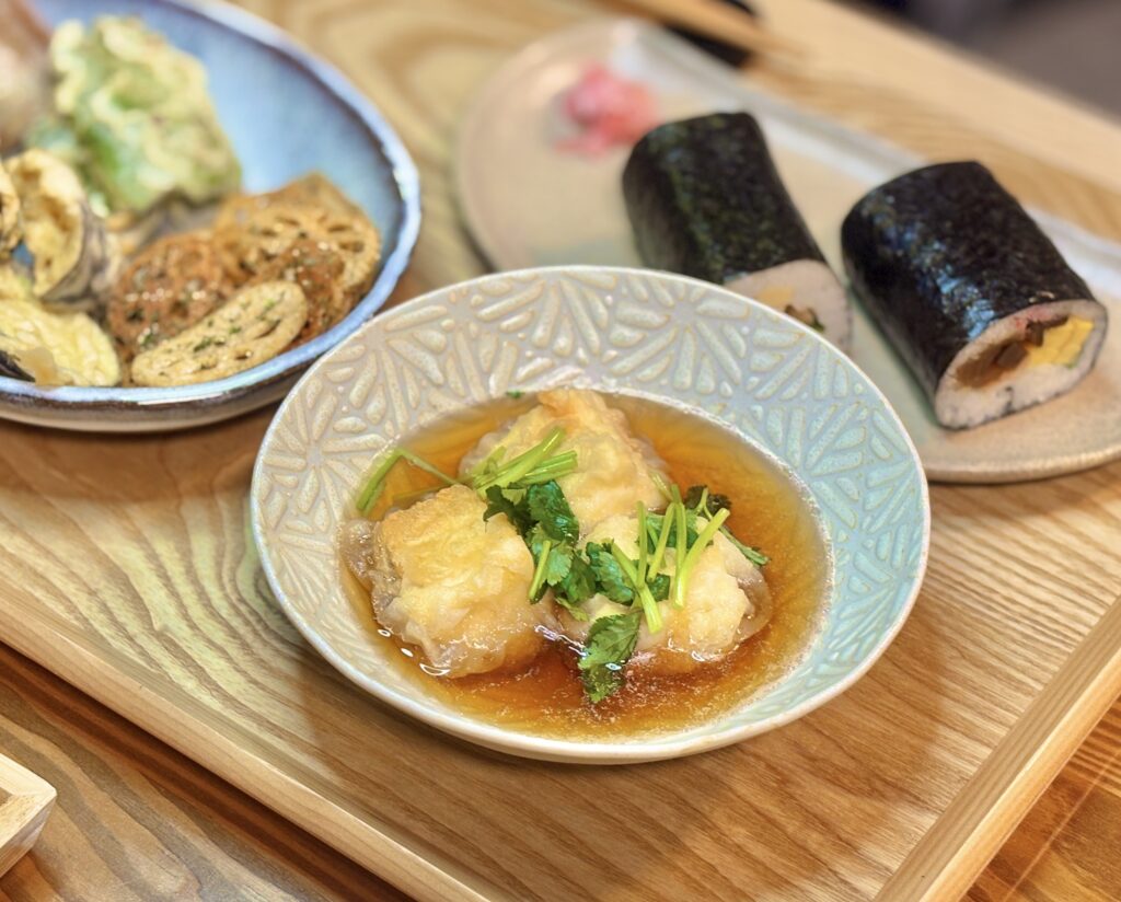 揚げ出し豆腐が器に盛られ、だしが張られている。豆腐の上には三つ葉がのせられ、温かみのある見た目。背景には太巻き寿司と野菜の揚げ物が並び、節分の食卓全体が感じられる。
