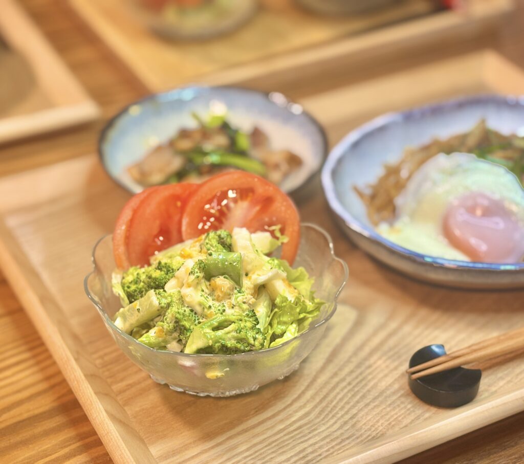 木製トレーに乗った定食を斜め前から撮影した写真で、手前のサラダに焦点が合っている。ガラスの器には、マヨネーズで和えたブロッコリー、ゆで卵、レタスが盛られ、その上にスライスされた赤いトマトが二切れ乗っている。奥には、目玉焼きが乗った焼きそばと、小皿料理が背景としてぼけて写っている。
