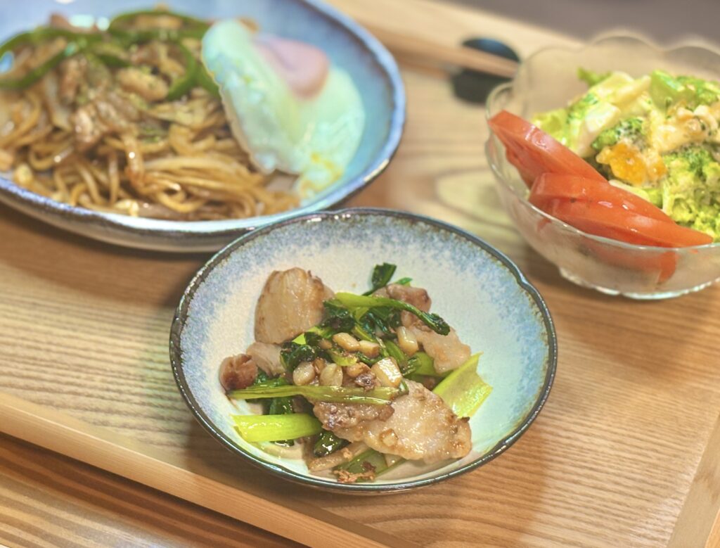 木製トレーの上の定食を斜め上から撮影したもので、手前にある小皿料理に焦点が合っている。青縁の小皿には、こんがりと炒められた豚肉と青菜が盛られており、具材の質感や焦げ目がはっきりと分かる。その奥には、目玉焼きが乗った焼きそばの大皿と、サラダが入ったガラスの器が背景としてぼけて写っている。
