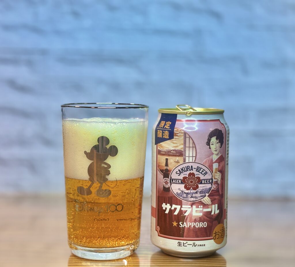 木製の表面の上に、サッポロビールの「サクラビール」の缶と、ビールが注がれたグラスが並んで置かれている。右側の缶は復刻版のデザインで、レトロな着物姿の女性がビールを持つイラストが描かれ、「限定醸造」の文字がある。左側のグラスにはミッキーマウスの全身シルエットと「Disney100」のロゴが入っており、黄金色のビールと白い泡で満たされている。背景は白いレンガ調の壁で、ぼかされている。
