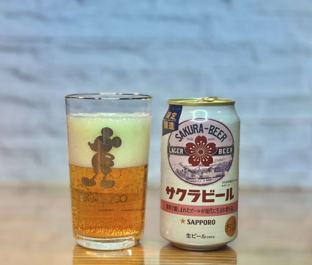 木製の表面の上に並んだ、サッポロ「サクラビール」の缶とビールが入ったグラス。右側の缶は、中央に大きな赤い桜のエンブレムと「SAKURA-BEER」の文字があり、その下にレトロなビール工場の風景が描かれたデザイン。「1913年頃のラベルをモチーフ」という説明書きが添えられている。左側のグラスはミッキーマウスのシルエット入りで、中身は泡立ったビールである。背景はぼかした白いレンガ調の壁。

