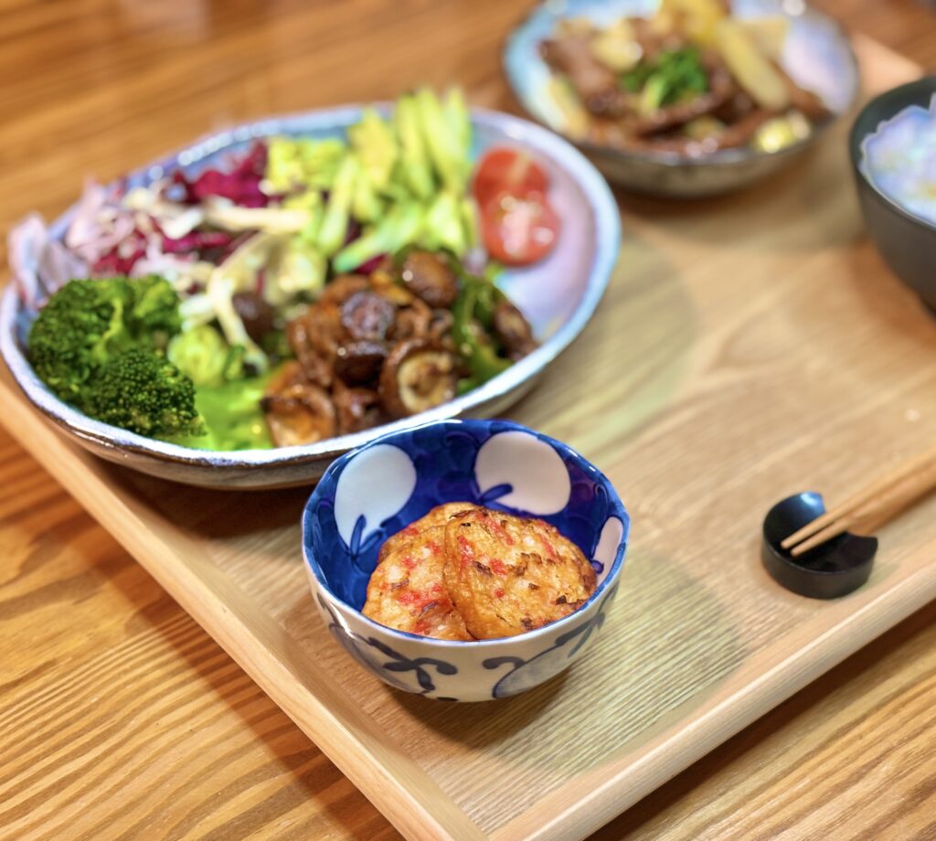木のトレーに並べた夕食の写真。手前の小鉢にかねてつの紅生姜天、奥の大皿には牛肉と椎茸とピーマンのオイスターソース炒めとブロッコリー、アボカド、紫キャベツなどの彩り野菜サラダが盛り付けられている。