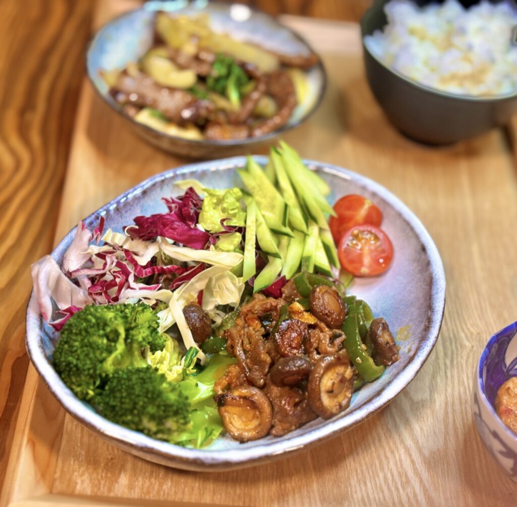 大皿に盛られた主菜とサラダのアップ。ブロッコリー、レタスや紫キャベツ、アボカド、ミニトマトとともに、牛肉と椎茸とピーマンのオイスターソース炒めが彩りよく盛り付けられている。