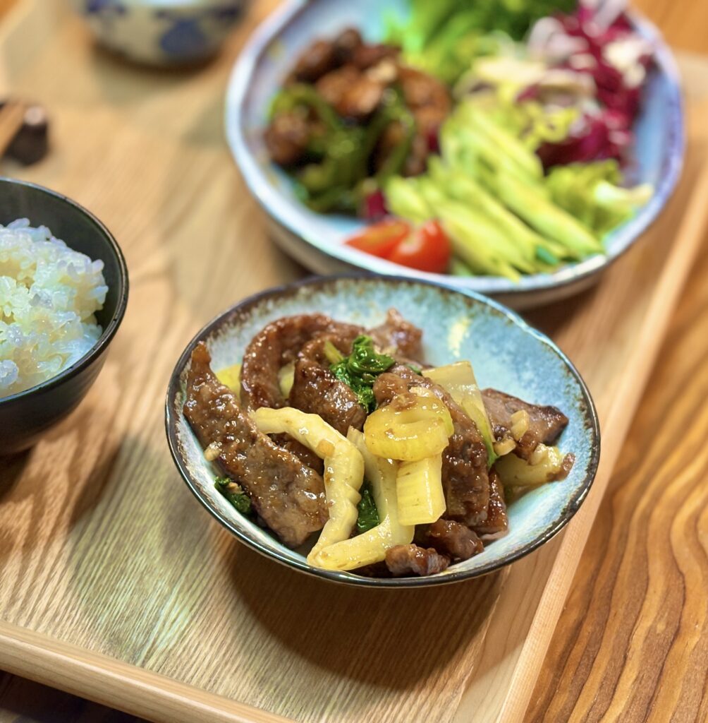 小鉢に盛られた牛肉とセロリの中華炒めの写真。照りのある牛肉とセロリが器に盛られ、奥にはサラダとご飯がぼけて写っている。
