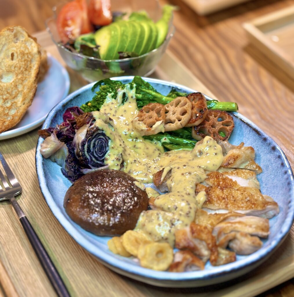 青い縁取りのある白い楕円皿に、美しく盛り付けられたチキンソテー。菜の花の蕾と茎がソースを纏い、シャキシャキとした蓮根の質感とコントラストをなしている。手前にはカリカリに焼かれたスライスニンニクが添えられ、食感の楽しさが伝わってくるような至近距離のカット。