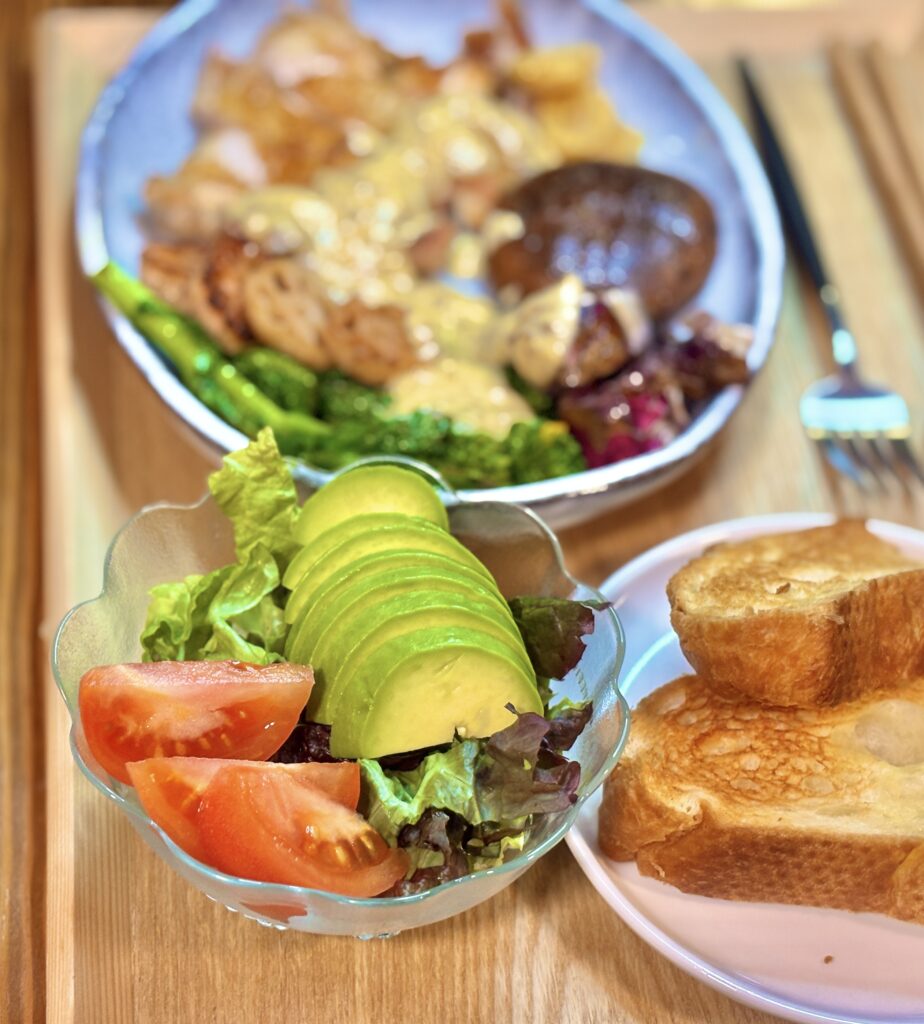 ガラスの器に丁寧に盛り付けられたサイドサラダ。完璧な厚さにスライスされ、扇状に並べられたアボカドが艶やかに輝いている。その横には真っ赤なトマトが添えられ、瑞々しさが際立つ。奥にはこんがり焼かれたトーストが配置され、ランチの満足感を高めてくれる新鮮な一品。