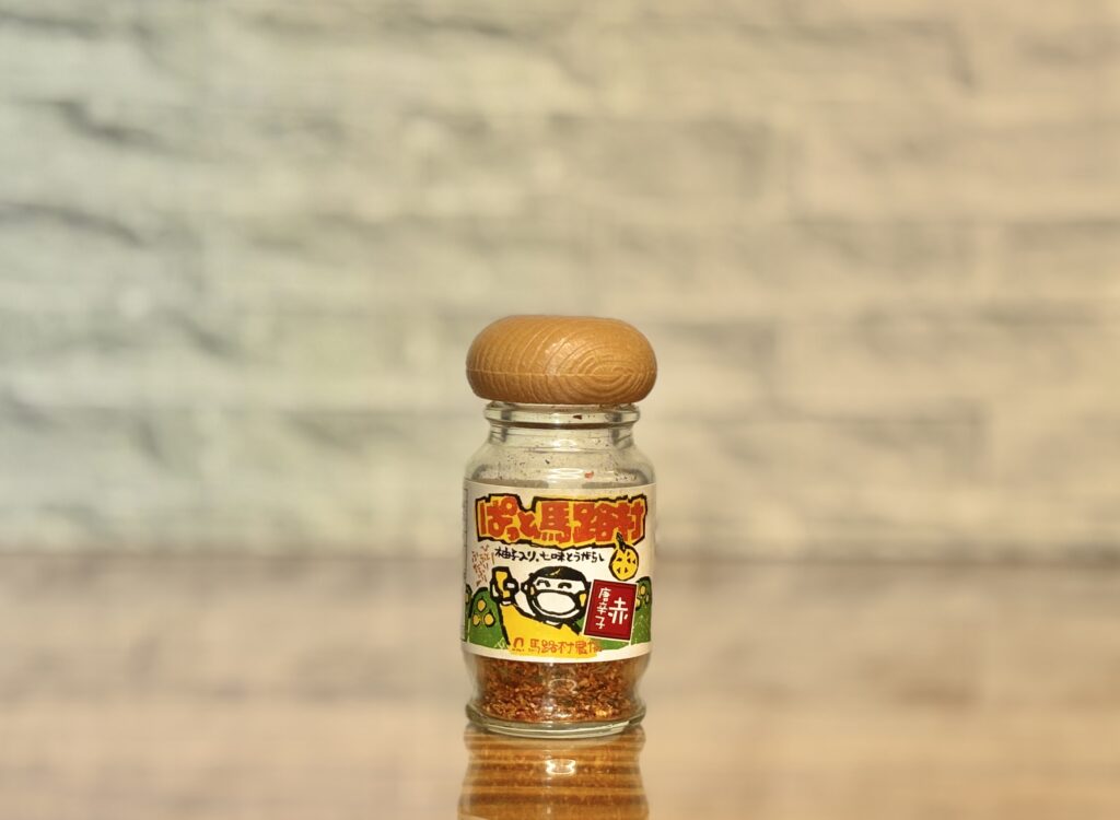 木製のフタが付いたガラス瓶に入った七味唐辛子を、テーブルの上に立てて撮影した写真。ラベルにはイラストと文字が描かれており、中の七味が見えるシンプルな構図で、料理のアクセントとして使われている様子が伝わる。
