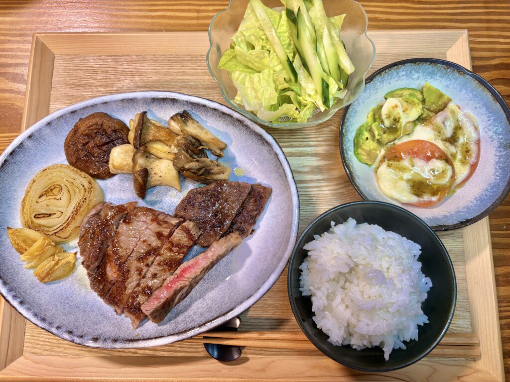 黒毛和牛ステーキときのこのガーリックソテー、玉ねぎステーキ、トマトとアボカドのモッツァレラチーズ焼き、サラダと白ごはんを木製トレーに盛り付けた夕食全体
