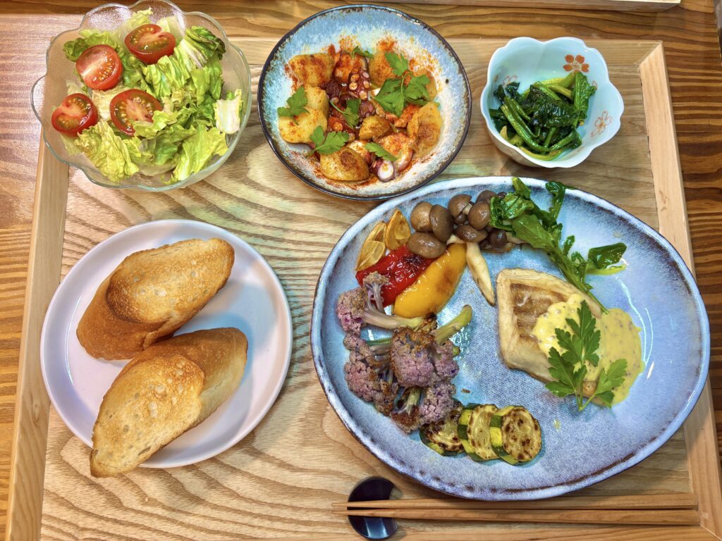 木製テーブルに並んだ洋風の食卓。大皿には真鯛のマスタードクリームソースが盛られ、クレソン、パプリカ、椎茸、カリフラワー、ズッキーニの焼き野菜が添えられている。奥にはタコのガリシア風、ほうれん草のガーリックソテー、ミニトマトとロメインレタスのサラダ、小皿の塩、スライスしたパンが並ぶ。オリーブオイルとワイングラスも置かれ、背景にはワインボトルが並ぶ棚が見える、落ち着いた雰囲気のダイニングシーン。