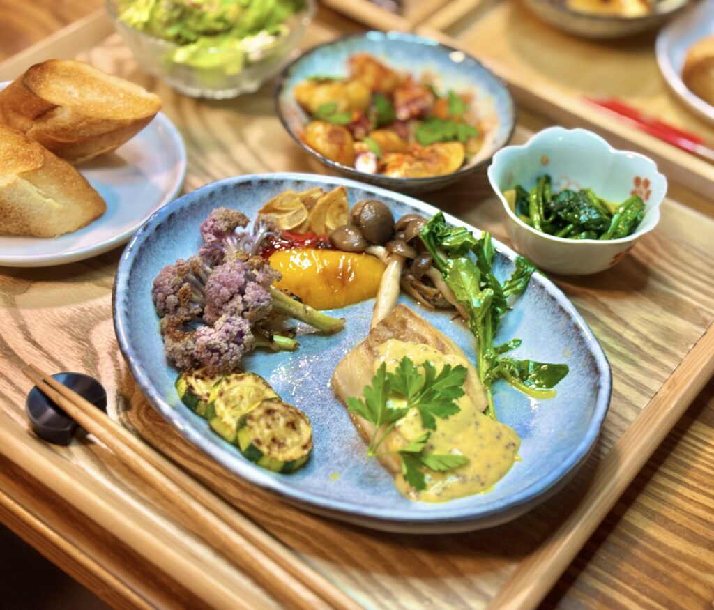 斜めから撮影されたメインプレートのアップ写真。マスタードクリームソースがかかった真鯛にイタリアンパセリが飾られ、手前には焼き色のついたズッキーニ。奥にはしめじ、パプリカ、紫カリフラワーが並び、立体感のある盛り付けが印象的。周囲の小鉢やパンはやわらかくぼけている。