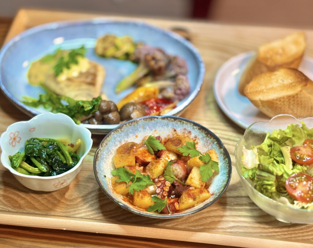 タコのガリシア風を中心にした写真。パプリカパウダーがまぶされたじゃがいもとタコが器に盛られ、緑のハーブが散らされている。手前には艶のあるほうれん草のガーリックソテー。奥に真鯛のプレートとパンがぼんやりと写り、温かみのある食卓の雰囲気が伝わる。