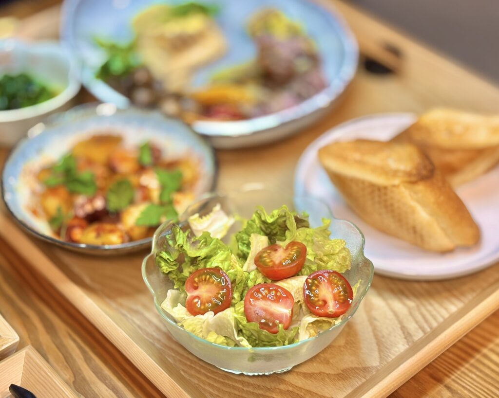 ロメインレタスとミニトマトのサラダのアップ写真。透明なガラスボウルに盛られ、半分にカットされたミニトマトの赤とレタスの淡い緑のコントラストが鮮やか。奥にはパンとメイン料理が背景にぼけて写っている。