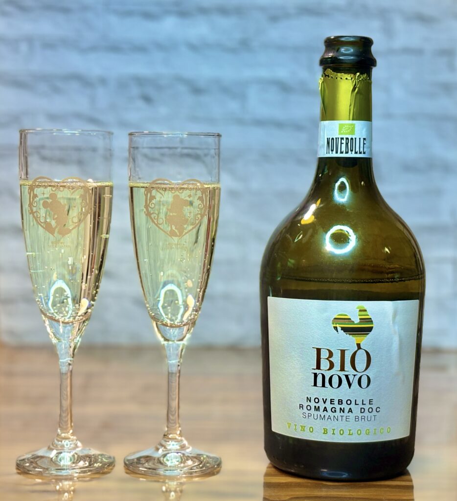 スパークリングワインのボトルとフルートグラス2脚の写真。淡い黄金色のワインが注がれ、細かな泡が立ち上っている。ボトルには「BIO novo」「Novebolle Romagna DOC Spumante Brut Vino Biologico」と記載されたラベルが見える。グラスにはハートとキャラクターの装飾が施され、落ち着いた背景の前に上品に置かれている。