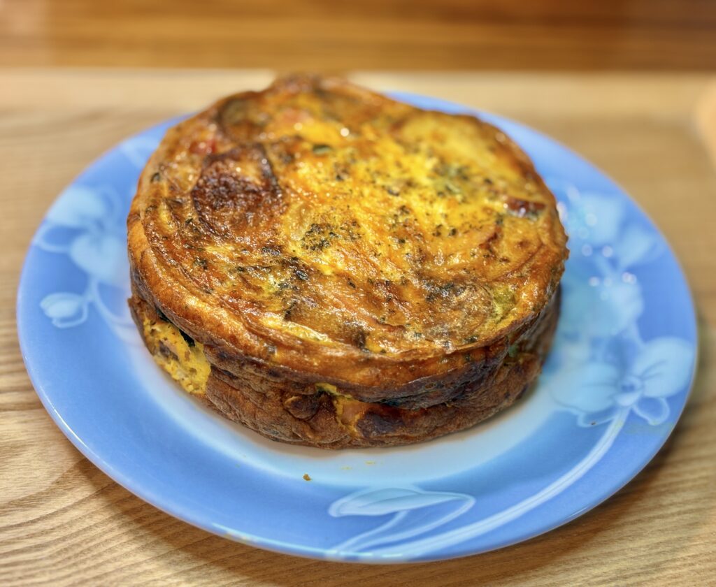 木目のテーブルの上に置かれた、淡いブルーのガラス皿に乗った丸ごと1個のスペインオムレツ。ケーキのように厚みを持たせて丸く焼かれており、表面はこんがりとした焼き色がついてふっくらと仕上がっている。