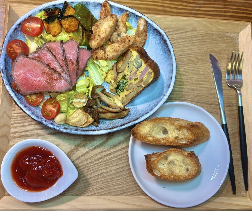 木製トレーの上に洋風のワンプレートとバゲットが並ぶ俯瞰写真。青みがかった大皿には、美しいピンク色の黒毛和牛ローストビーフ、分厚いスペインオムレツ、キツネ色に揚がった鳥ささみ揚げが盛られ、かぼちゃ、グリーンパプリカ、しめじの焼き野菜と、フライドガーリック、ミニトマト、レタスが彩りよく添えられている。右下にはこんがり焼いたバゲットが2切れ乗った白い小皿、左下にはケチャップが入った白い小鉢、右側には黒い柄のナイフとフォークが置かれている。