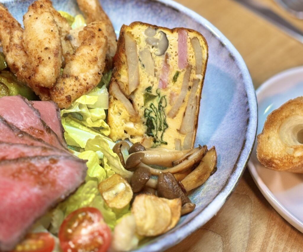 洋風ワンプレートのクローズアップ写真。手前には具だくさんな極厚スペインオムレツの断面が見え、中にじゃがいも、ほうれん草、しめじ、ベーコンがぎっしり詰まっている様子がわかる。その後ろや横には、ローストビーフ、サクサクの鳥ささみ揚げ、焼き野菜などが所狭しと並べられている。