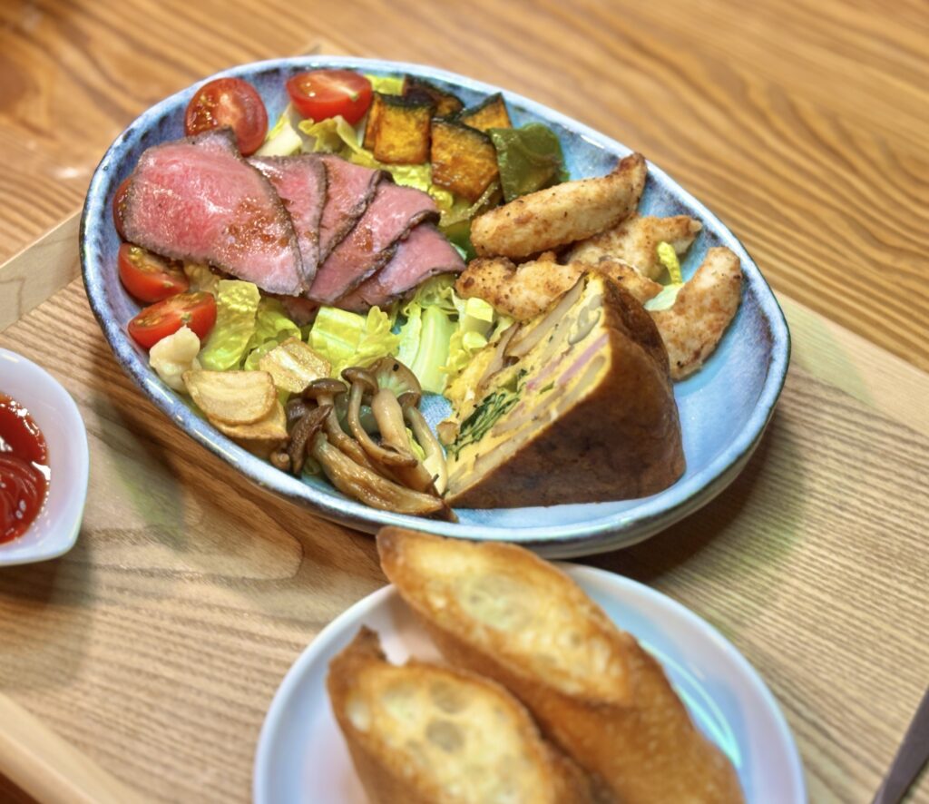 木製トレーの上に洋風のワンプレートとバゲットが並ぶ俯瞰写真。青みがかった大皿には、美しいピンク色の黒毛和牛ローストビーフ、分厚いスペインオムレツ、キツネ色に揚がった鳥ささみ揚げが盛られ、かぼちゃ、グリーンパプリカ、しめじの焼き野菜と、フライドガーリック、ミニトマト、レタスが彩りよく添えられている。右下にはこんがり焼いたバゲットが2切れ乗った白い小皿、左下にはケチャップが入った白い小鉢、右側には黒い柄のナイフとフォークが置かれている。