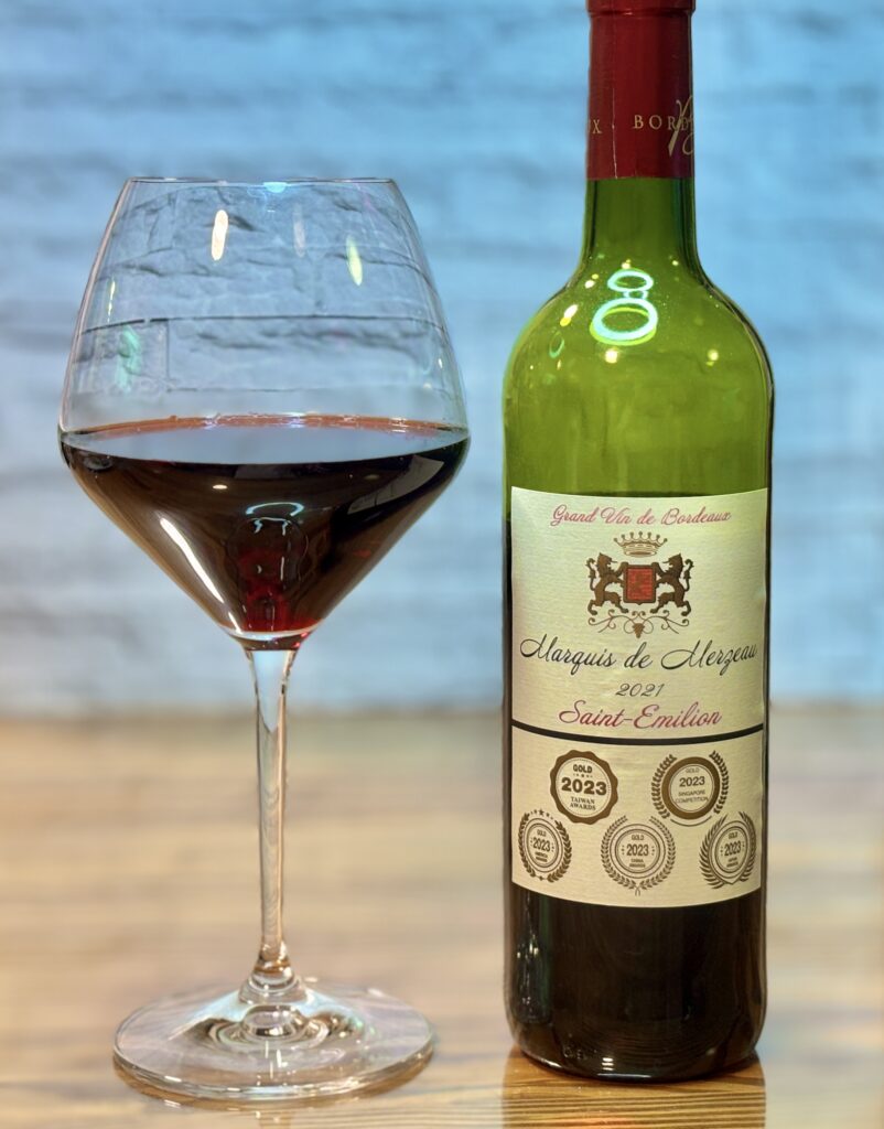 木目のテーブルの上に置かれた赤ワインのボトルと、赤ワインが注がれた大きなワイングラス。ボトルは「マルキ・ド・メルゾー サン・テミリオン(Marquis de Merzeau Saint-Emilion 2021)」で、ラベルには複数の金賞メダルがデザインされている。背景には白いレンガ調の壁が見える。