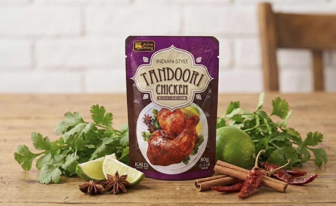 インド風タンドリーチキンのレトルトパウチ商品が木製テーブルの上に立てて置かれている。パッケージは紫色で、中央に焼き色のついたチキンの写真があり、「TANDOORI CHICKEN」と書かれている。周囲にはパクチー、ライム、シナモンスティック、スターアニス、赤唐辛子などのスパイスやハーブが飾られている。
