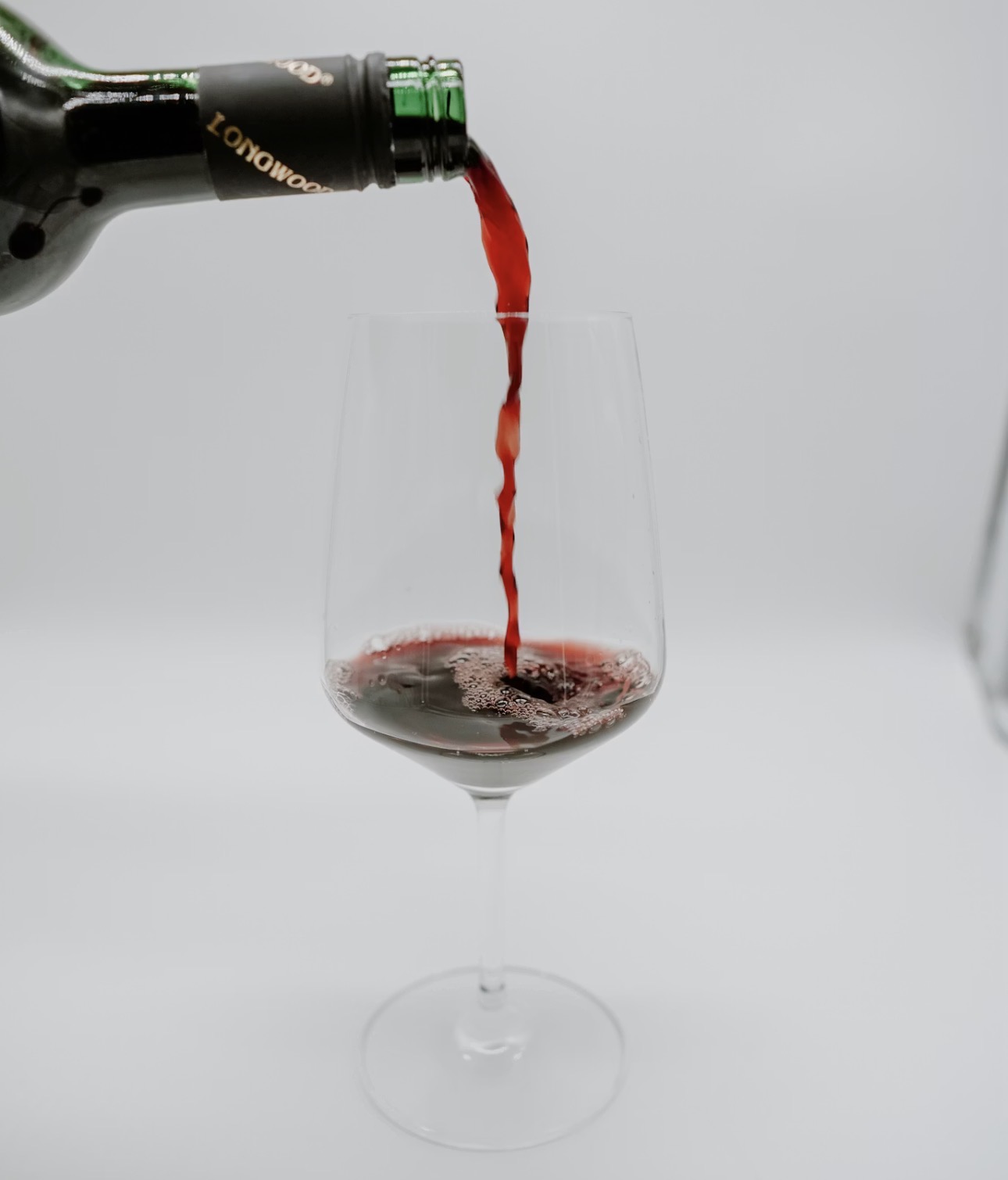 ワインボトルから赤ワインが注がれ、透明なワイングラスに赤い液体が流れ込んでいる様子。背景は白でシンプルな構図。