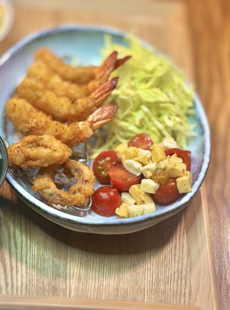 エビフライと副菜をクローズアップした一枚。網にのせられたエビフライの衣の粒立ちがはっきり見え、横にはリング状のフライ。たっぷりの千切りキャベツの隣に、ミニトマトと角切りのチェダーチーズ、モッツァレラチーズを和えたマリネが鮮やかに並ぶ。木目のトレーが背景となり、揚げたての香ばしさとみずみずしい副菜の対比が伝わる。