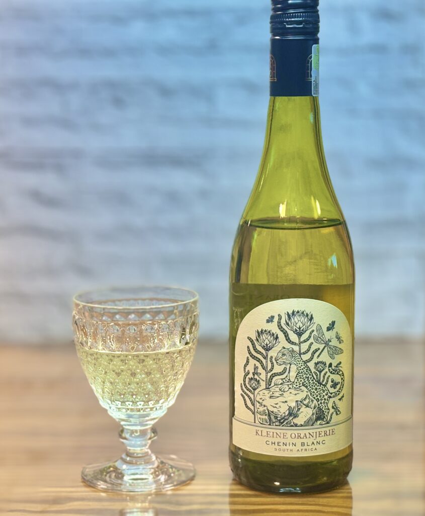 テーブルの上に、白ワインのボトルと模様入りのグラスを並べた写真。ボトルのラベルにイラストがあり、「KLEINE ORANJERIE CHENIN BLANC SOUTH AFRICA」と読める