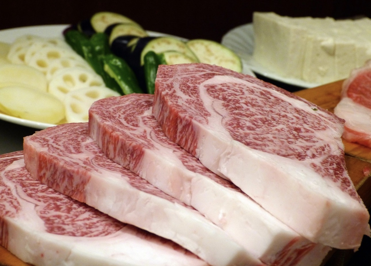 霜降りが美しく入った厚切りの和牛ステーキ肉が数枚重ねられ、奥にはれんこん、なす、ししとう、豆腐などの野菜が並んでいる様子。