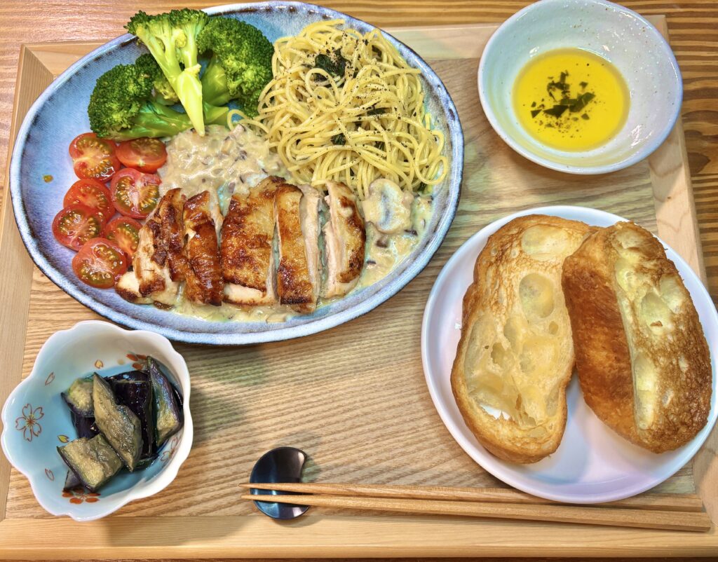 木のトレイに美しくセットされた和洋折衷のおうちごはん。中央の青みがかった大きなお皿には、こんがりと焼き色のついたグリルチキンが並び、たっぷりのキノコクリームソースがかけられています。同じお皿には、バジルパスタ、鮮やかな緑のブロッコリー、半分にカットされた赤いミニトマトが彩りよく添えられています。左下には花びら型の小鉢に入った茄子の煮浸し、右側にはこんがり焼けたバゲット2枚、ハーブが浮かんだオリーブオイルの小鉢、そして手前には箸置きにのせられた木のお箸が丁寧に並べられています。温かみのある食卓の風景です。

