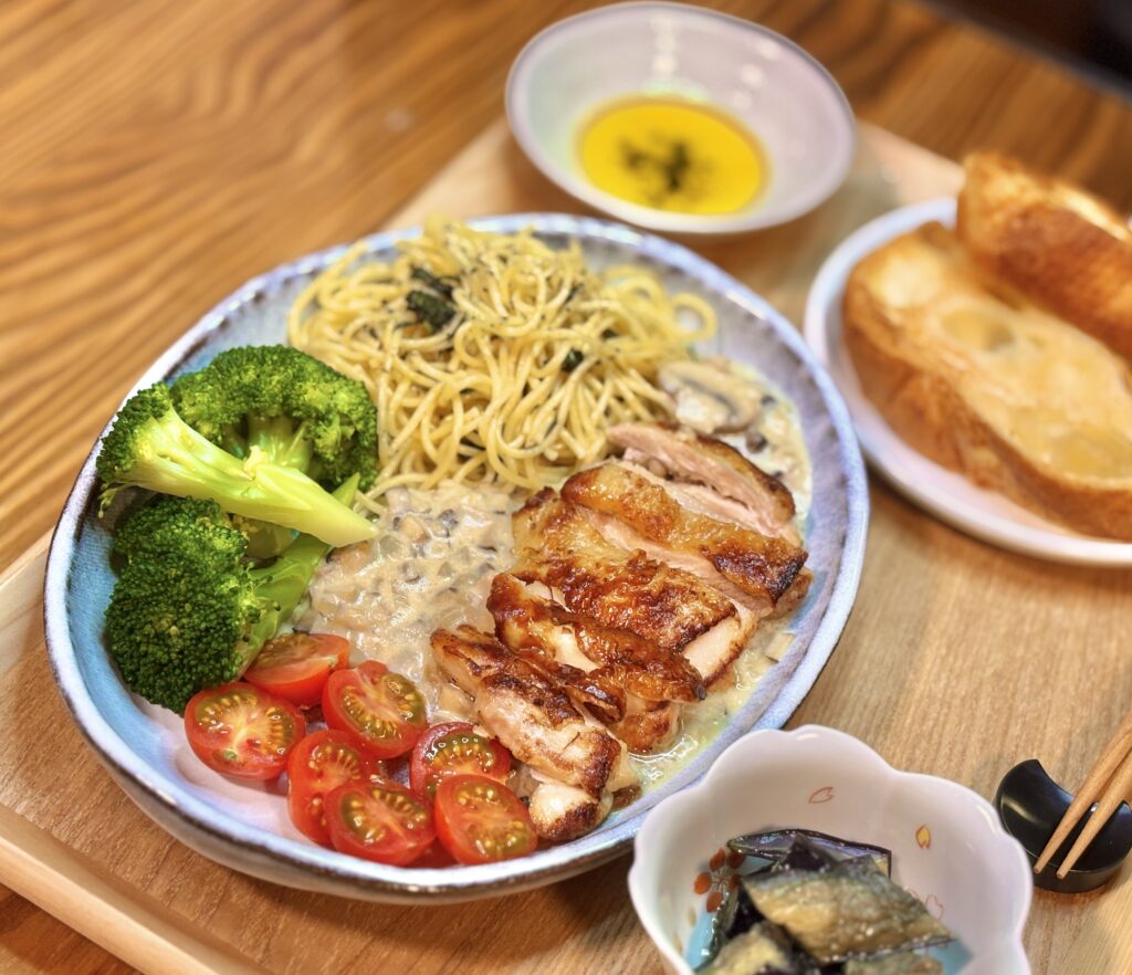 木製トレーの上に、こんがり焼いたグリルチキンのスライスとマッシュルーム・椎茸のクリームソース、バジルパスタ、茹でブロッコリー、半分に切ったミニトマトを盛り付けたワンプレート。奥に黄色いスープ、右にトースト、小鉢に茄子の煮浸しが添えられている
