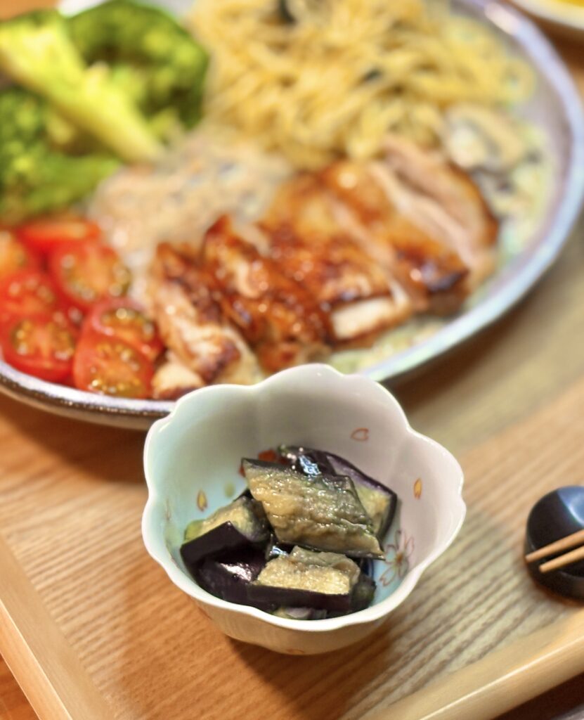 花形の小鉢に盛った茄子の煮浸しのアップ。奥にぼけたワンプレート（グリルチキンのクリームソース、バジルパスタ、ブロッコリー、ミニトマト）が写り、木製トレーと箸置きが見える