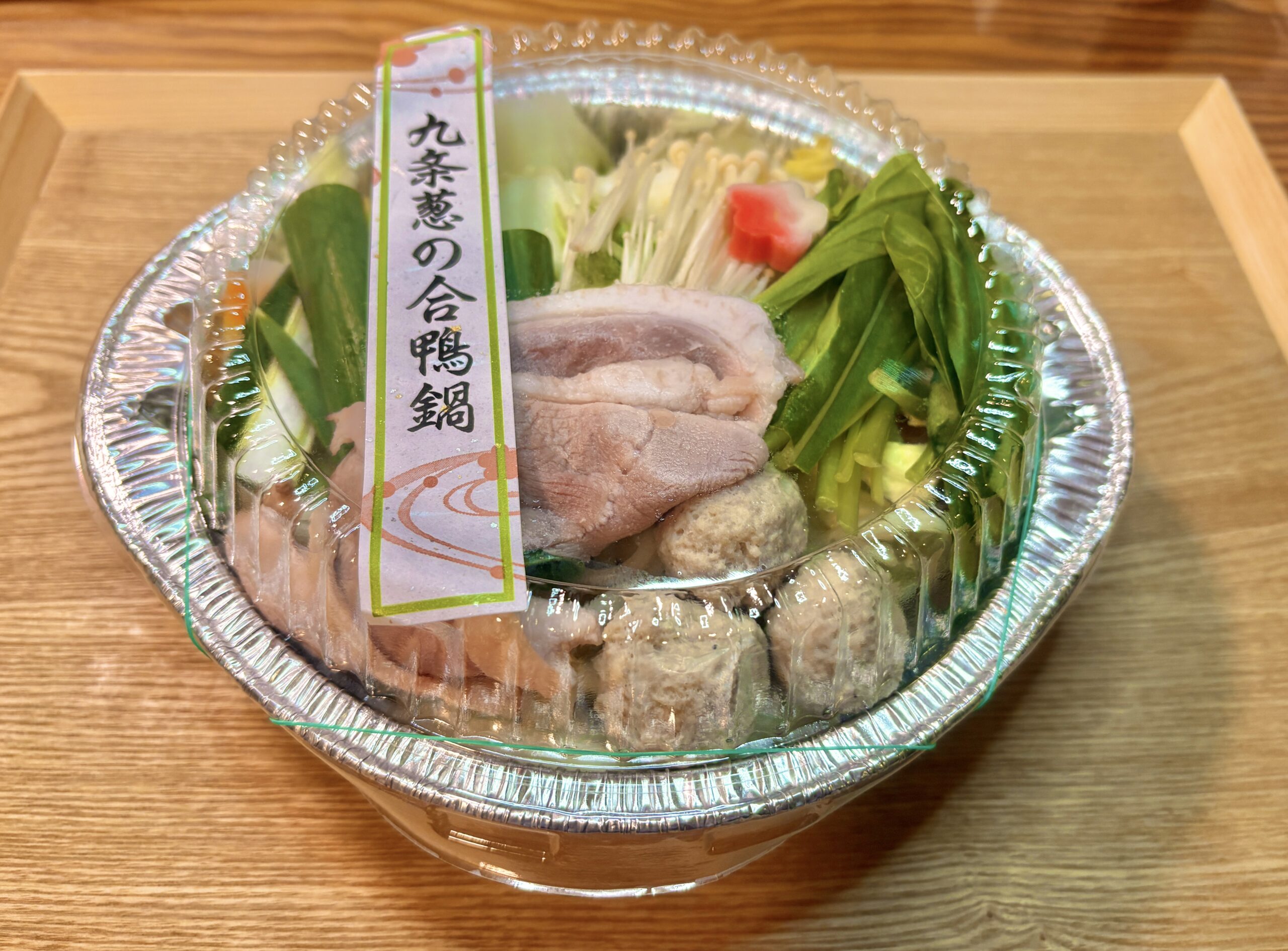 透明のふた付きアルミ鍋に「九条葱の合鴨鍋」のラベル。合鴨肉、九条ねぎ、えのき、白菜などの具材がきれいに詰められた鍋セットのアップ。