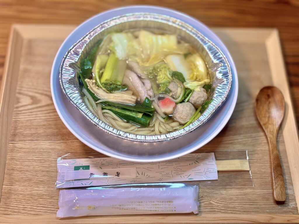 煮込んだ九条葱の合鴨鍋をアルミ鍋のまま器にのせた食卓写真。白菜や九条ねぎ、きのこ、つくね、合鴨肉がスープに浮かび、手前に割り箸とスープの小袋、右に木のスプーンが置かれている。