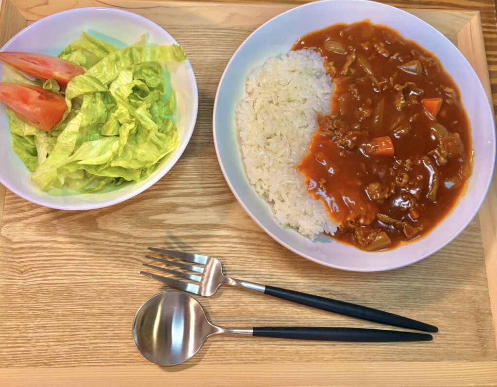 木製トレーの上に、白い器に盛られたハヤシライス。右半分にとろみのあるルーがかかり、牛肉や玉ねぎ、にんじんが見える。左側にはレタスとトマトのシンプルなサラダの小鉢。手前には黒い持ち手のスプーンとフォークが並べられている。