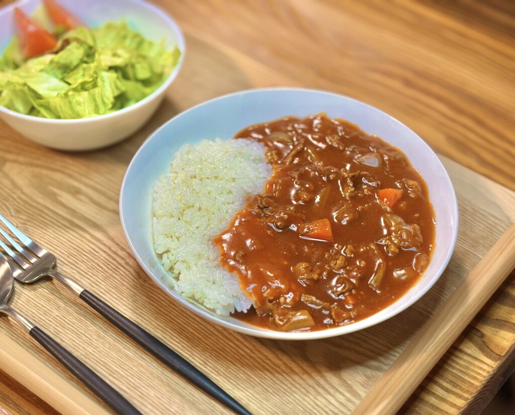 斜めから撮影したハヤシライスのプレート。白い皿にごはんととろみのある赤いソースが半分ずつ盛られ、牛肉や野菜の具材が確認できる。奥にはレタスとトマトのサラダがぼんやり写り、木目のトレーが温かみのある食卓を演出している。