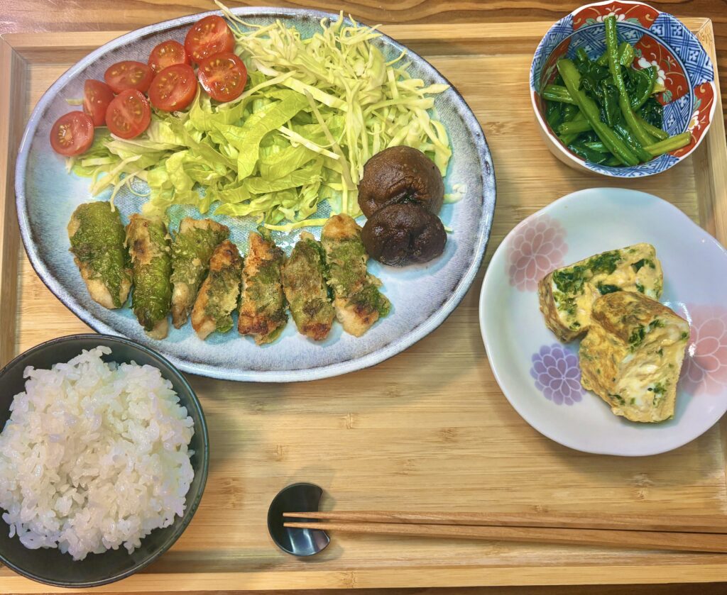 木製トレーに並べられた和定食の全体写真。青い大皿には鶏胸肉の紫蘇巻きのこんがり焼き、千切りキャベツとレタス、半分に切ったミニトマト、焼き椎茸が盛られている。小皿には青さ入りの卵焼き、別の小鉢には青菜のおひたし。黒い茶碗には白ごはんがよそわれ、箸が添えられている。