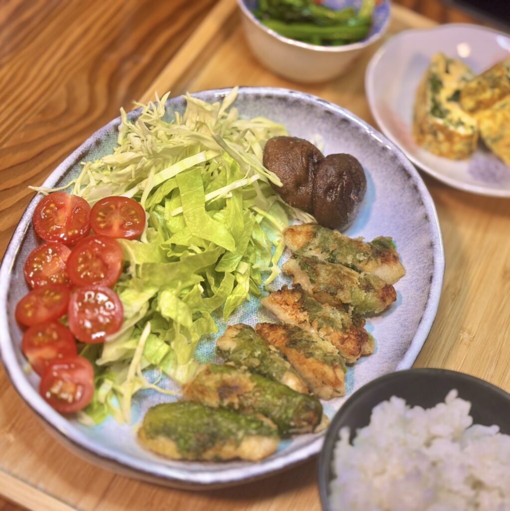 青い大皿を中心にした料理のアップ写真。こんがり焼き色のついた鶏胸肉の紫蘇巻きが並び、横には焼き椎茸。みずみずしいレタスと千切りキャベツ、艶のあるミニトマトが彩りを添えている。手前には白ごはんの茶碗が見える。
