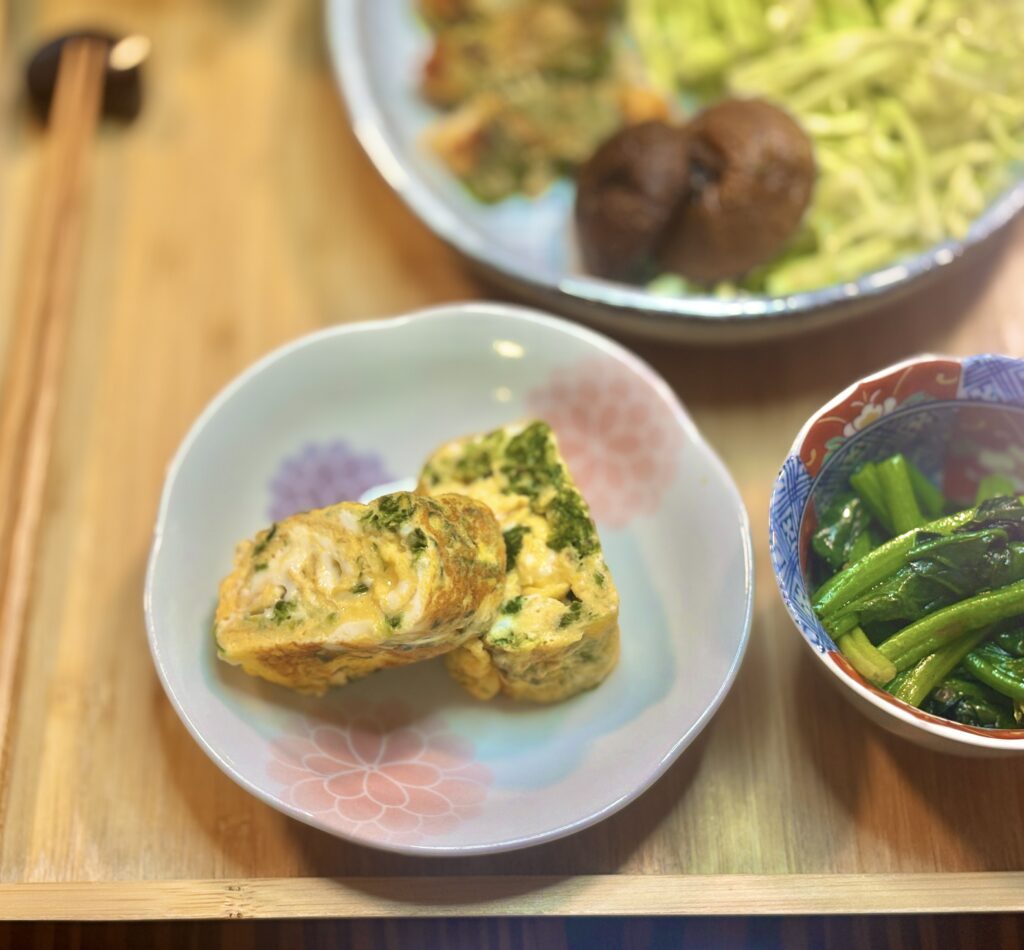 青さ入りの卵焼きのアップ写真。ふんわりと巻かれた卵焼きを切り分けた断面に、緑の青さが散りばめられている。隣には青菜のおひたしの小鉢が置かれ、背景には大皿の料理がぼんやりと写っている。
