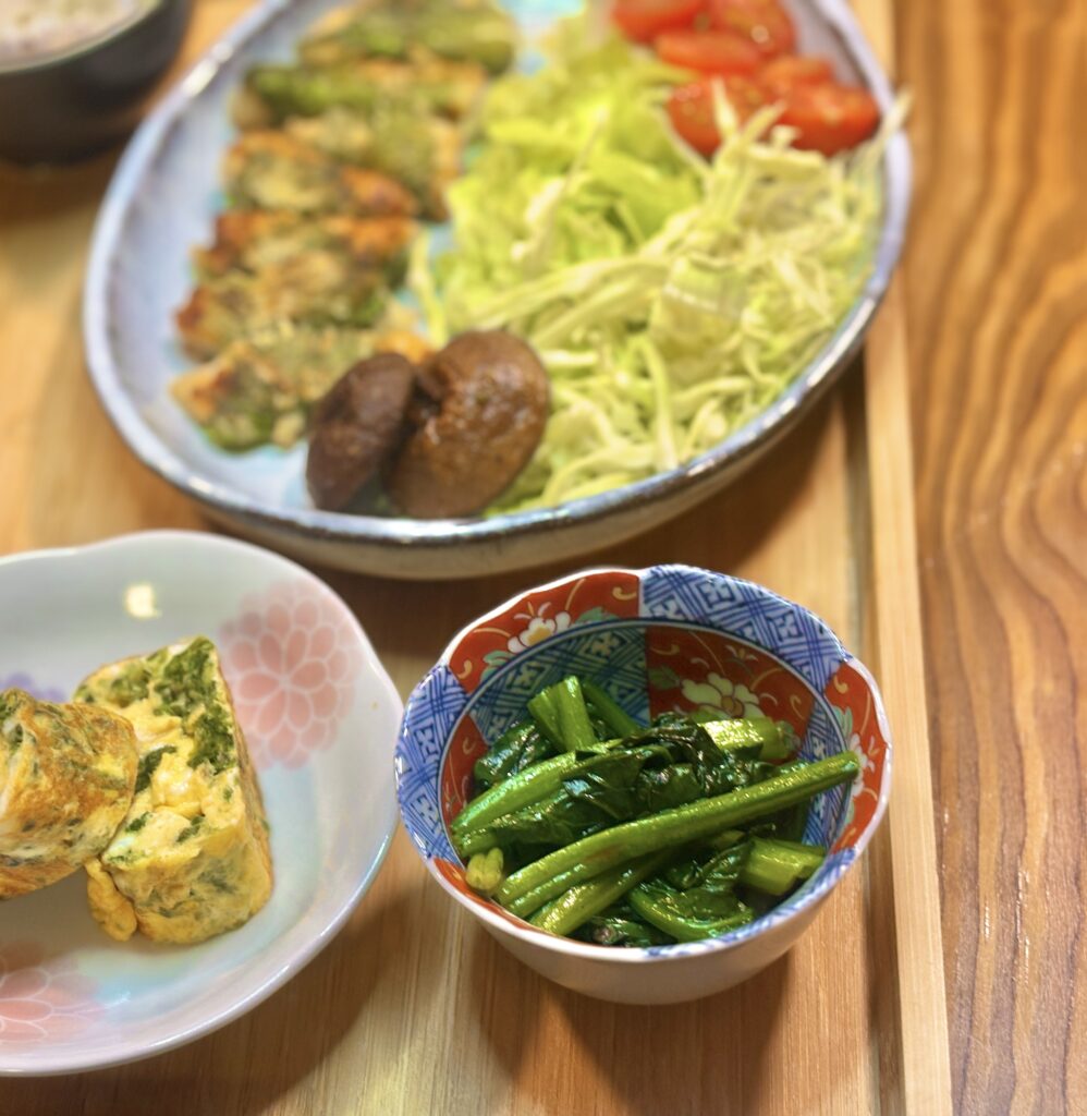 
小鉢に盛られた青菜のおひたしのアップ写真。鮮やかな緑色の青菜がつややかに和えられている。奥には鶏胸肉の紫蘇巻きやサラダ、焼き椎茸がのった大皿が見える。
