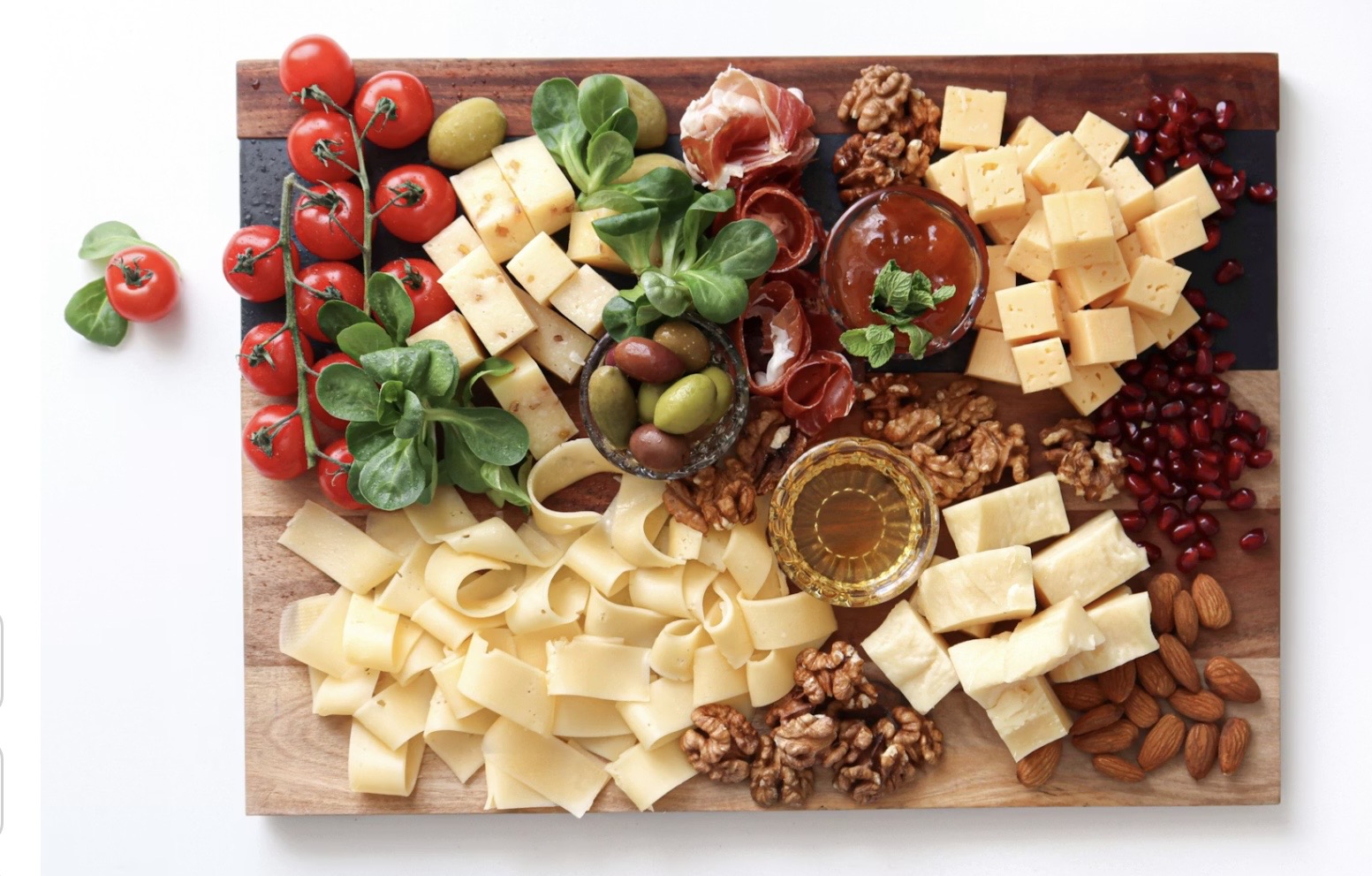 木製ボードに盛り付けられたチーズと前菜の盛り合わせ。角切りやスライス状のチーズ数種類、くるみとアーモンド、グリーンとブラックのオリーブ、生ハム、ミニトマト、ベビーリーフ、ザクロの実、はちみつやジャムの小鉢が彩りよく並んでいる。