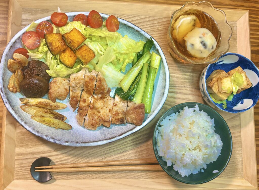 木のトレーの上に、カットした豚肉のソテーを盛った大皿と白ごはん、小鉢2品を並べた定食の全体写真。大皿にはレタスとミニトマト、かぼちゃ、青菜、しいたけ、焼きネギなどの副菜も彩りよく添えられている