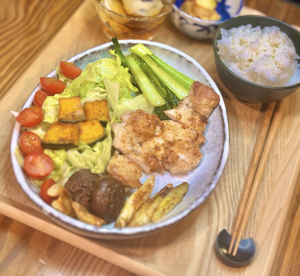豚肉のソテーを食べやすく切って盛り、レタスとミニトマト、かぼちゃ、青菜、しいたけ、焼きネギをワンプレートにまとめた一皿。奥に白ごはんと小鉢がぼんやり写り、温かい食卓の雰囲気が伝わる。