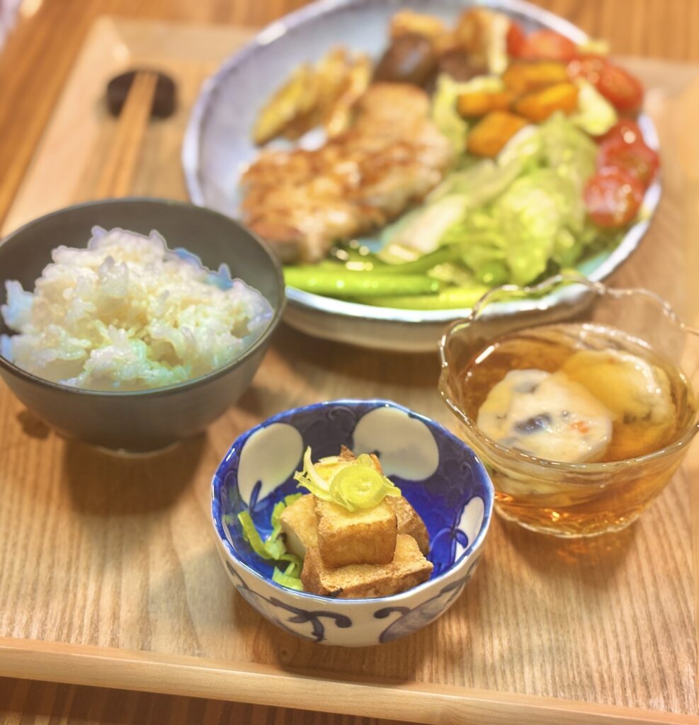 白ごはんの茶碗を手前に置き、奥に豚肉のソテーとたっぷり野菜のワンプレートが見える定食風景。手前には青い小鉢（厚揚げの角切りに薬味をのせたもの）と、琥珀色の汁物風の小鉢も添えられている。